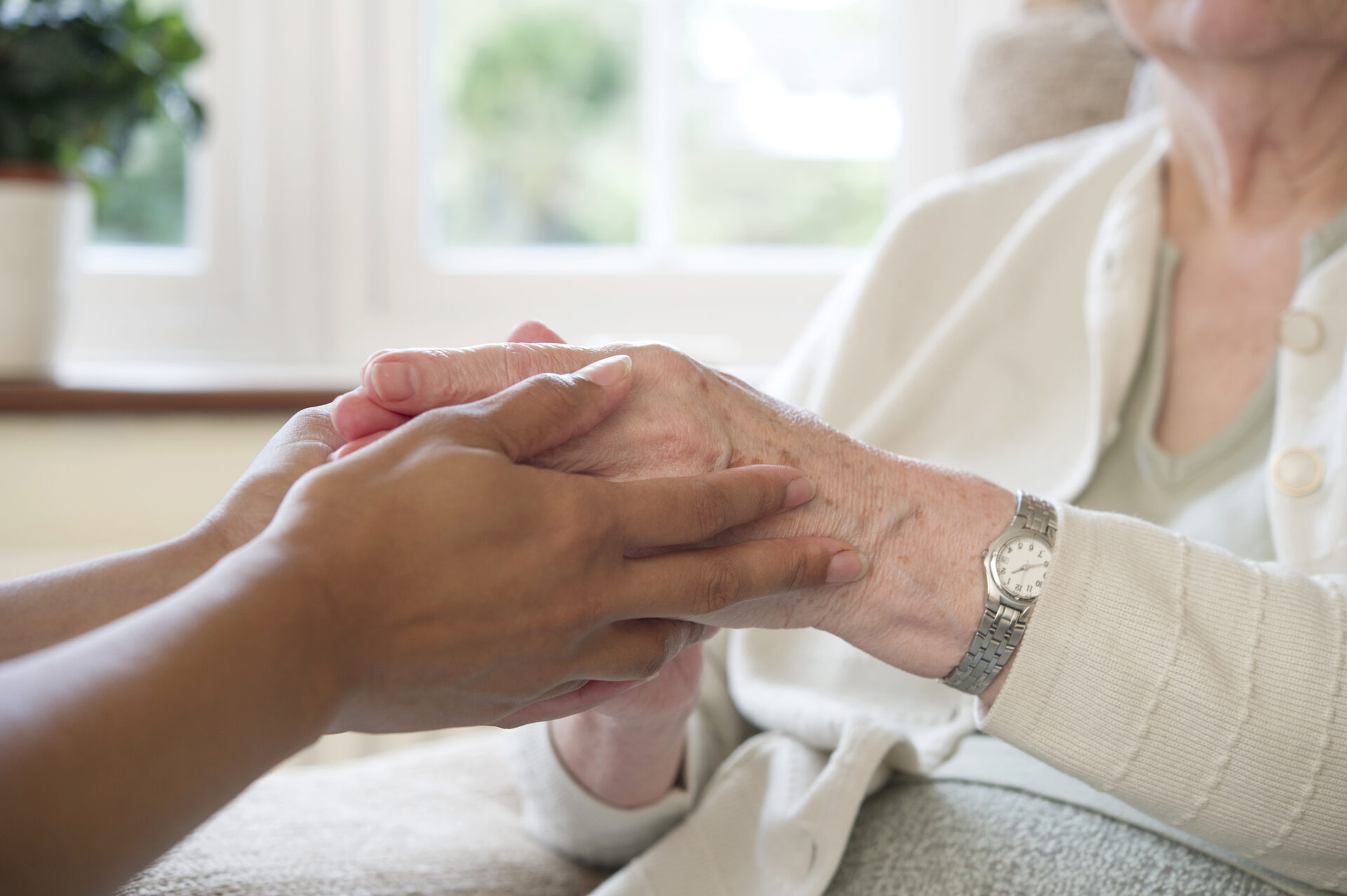 A carer holding the hands of an older woman sitting in a bright living room